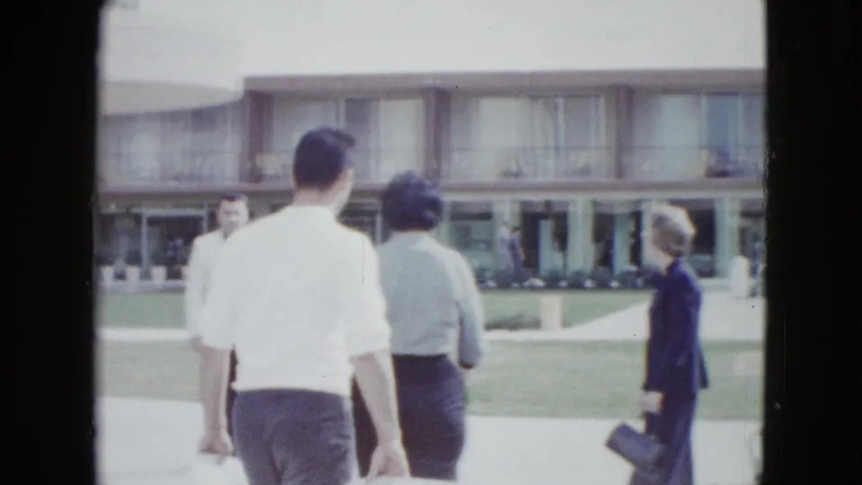Adults pass through the grounds of a mid-century Las Vegas motel, with a two-story facade and lawn behind them.