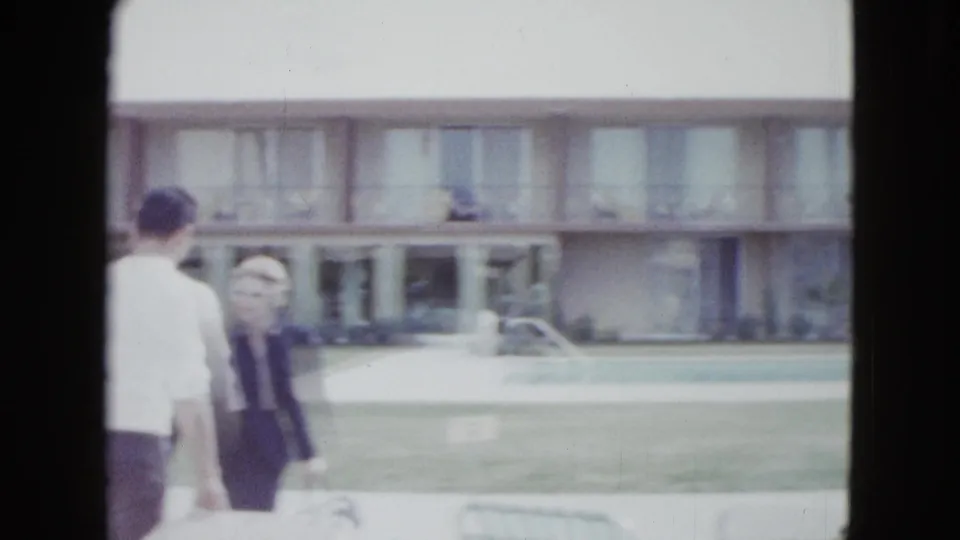 A blurred woman faces the camera while other adults walk beside a lawn and motel building in 1958 Las Vegas footage.