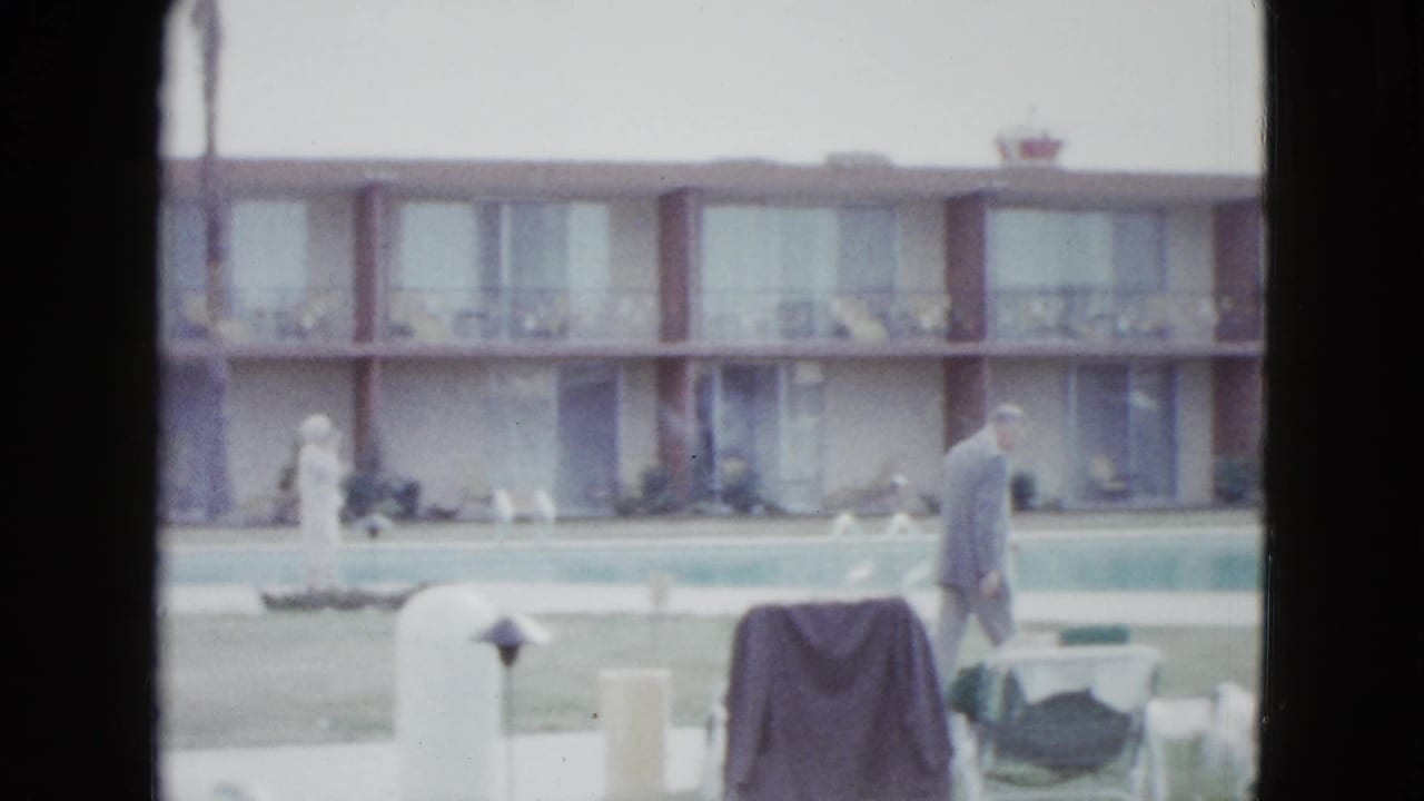 A man in a light-colored suit walks along a Las Vegas motel poolside with lounge chairs, lawn, and room balconies behind him.