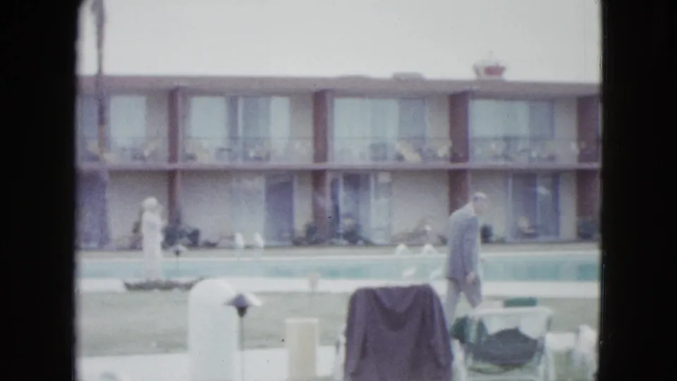 A man in a light-colored suit walks along a Las Vegas motel poolside with lounge chairs, lawn, and room balconies behind him.