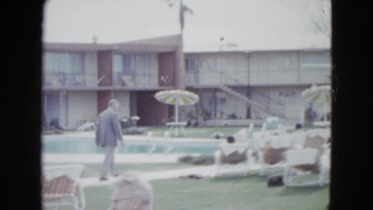 A man in a light suit walks near a Las Vegas motel pool as seated adults look toward the water, with balconies and palms behind them.