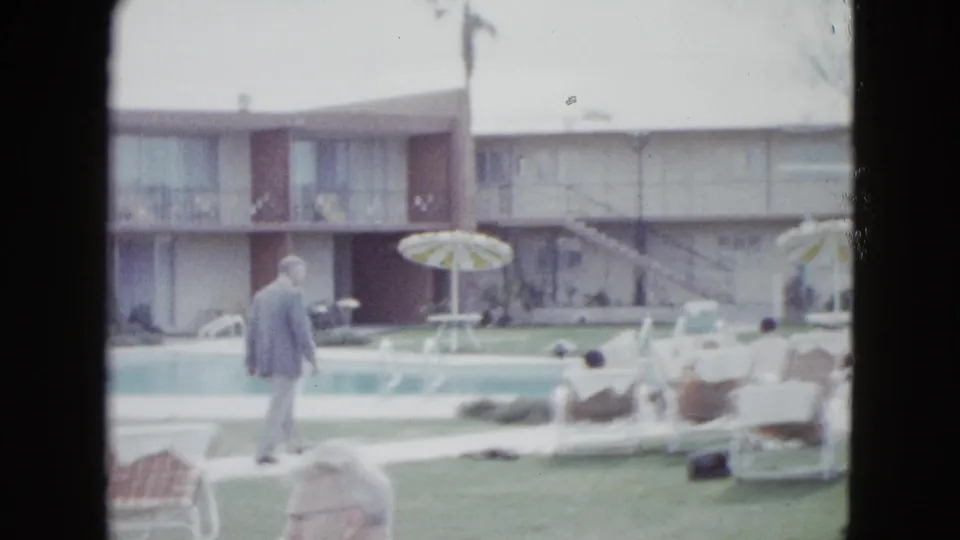 A man in a light suit walks near a Las Vegas motel pool as seated adults look toward the water, with balconies and palms behind them.