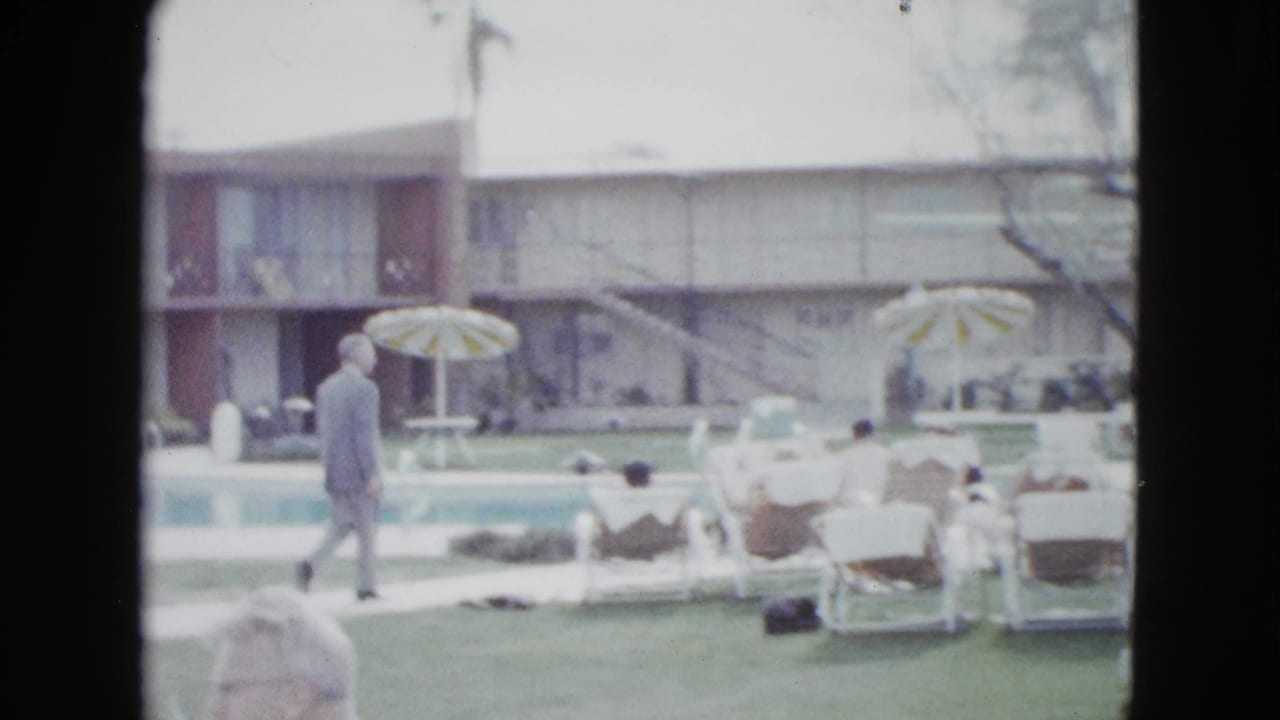 Guests relax near a hotel pool while a man walks along the lawn beside mid-century motel buildings.