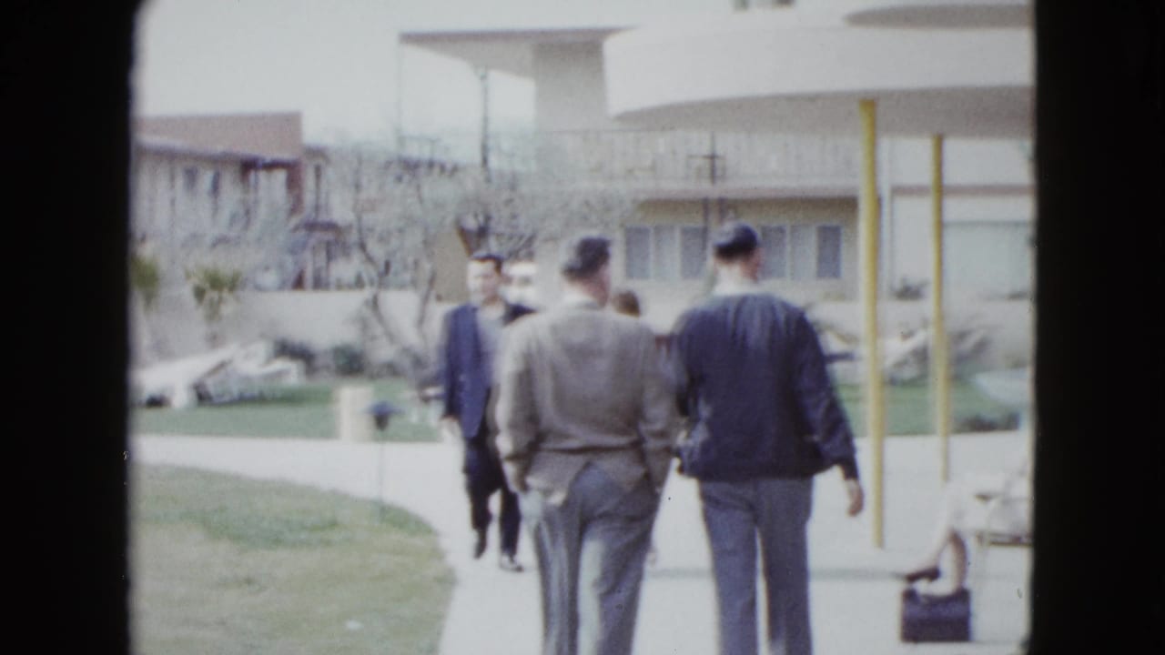 Men walk along a paved path beside lawn and motel buildings, with poolside seating in the background.
