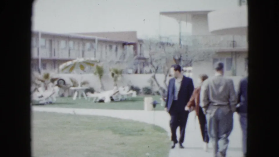 1958 Las Vegas hotel walkway scene with guests in suits and casual clothes moving past poolside furniture and motel buildings.