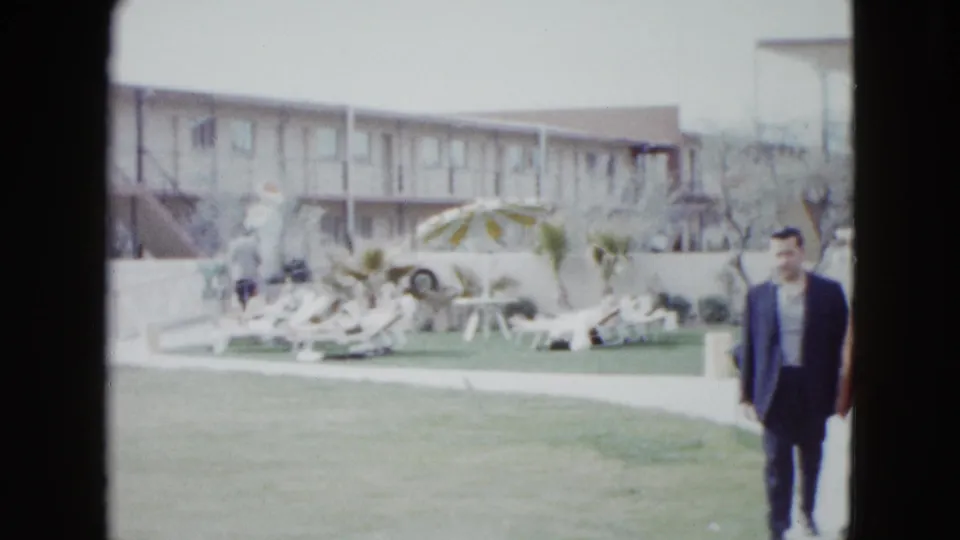 1958 Las Vegas motel courtyard with a man in the foreground, poolside loungers, striped umbrella, lawn, and two-story lodging behind.