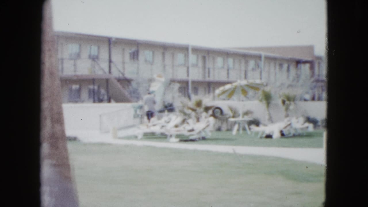 1958 Las Vegas home-movie frame of a motel pool area seen past palm trunks, with lawn, lounge chairs, umbrella, and a low building.