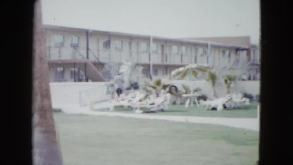 1958 Las Vegas home-movie frame of a motel pool area seen past palm trunks, with lawn, lounge chairs, umbrella, and a low building.