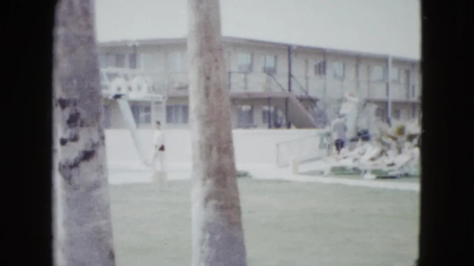 1958 Las Vegas resort scene with palm trunks in the foreground, a motel pool slide, lawn, white fence, and small figures near the pool.