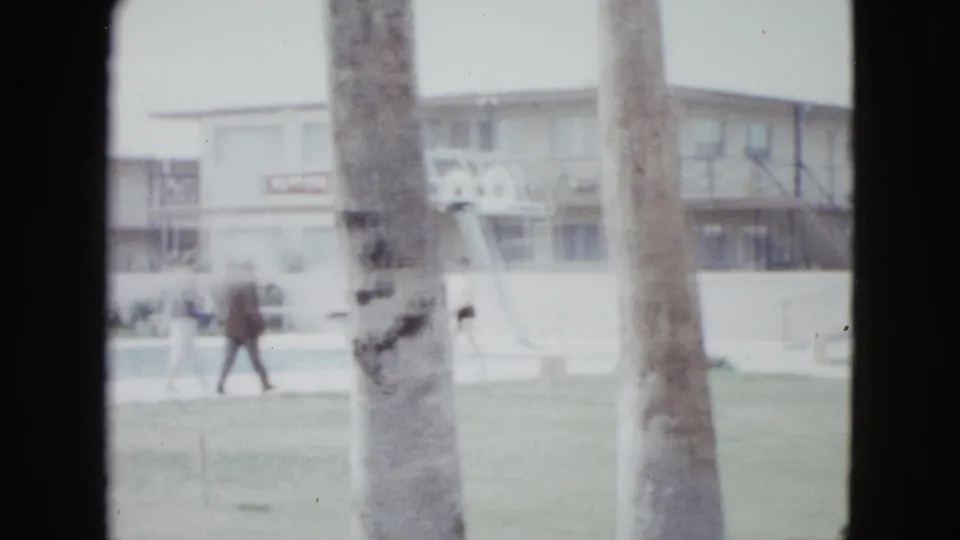 1958 Las Vegas home-movie frame of a guest walking across the lawn near palm trunks, with motel rooms and poolside features behind.