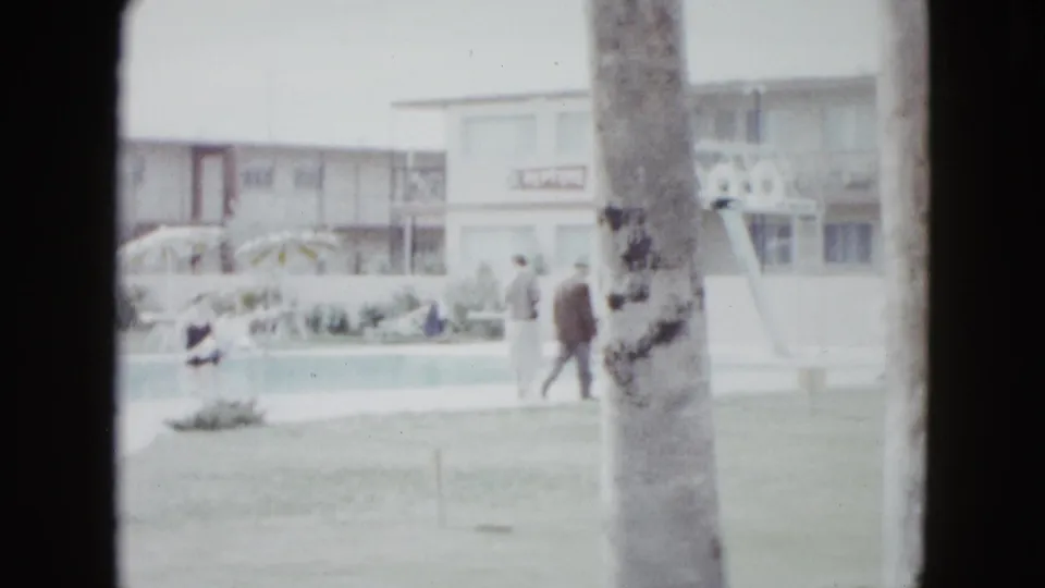 1958 Las Vegas poolside view with two guests walking near the lawn, palm trunks in front, and a low motel building behind.