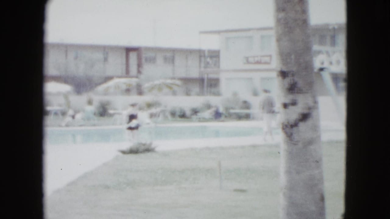 1958 Las Vegas resort pool area seen from a home-movie viewpoint, with lawn, palm tree, umbrellas, motel facade, and distant guests.