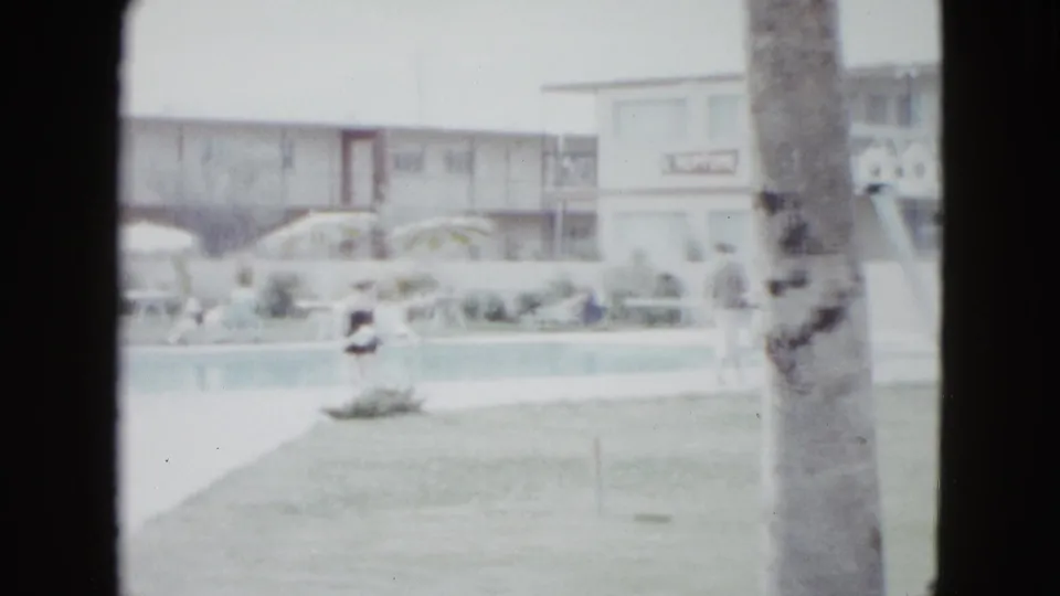 1958 Las Vegas resort pool area seen from a home-movie viewpoint, with lawn, palm tree, umbrellas, motel facade, and distant guests.