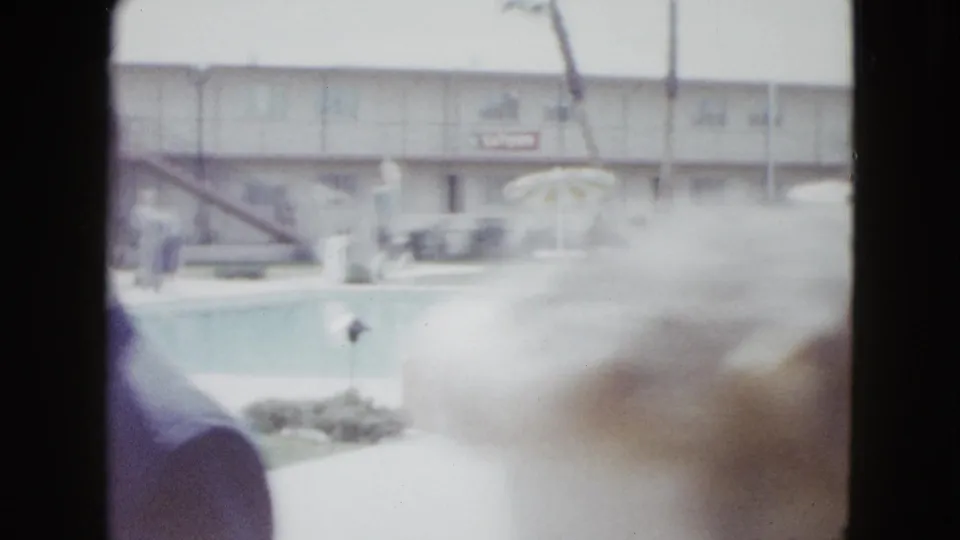 Soft 1958 Las Vegas poolside frame with motel facade, umbrellas, guests by the water, and a blurred person passing close to the camera.