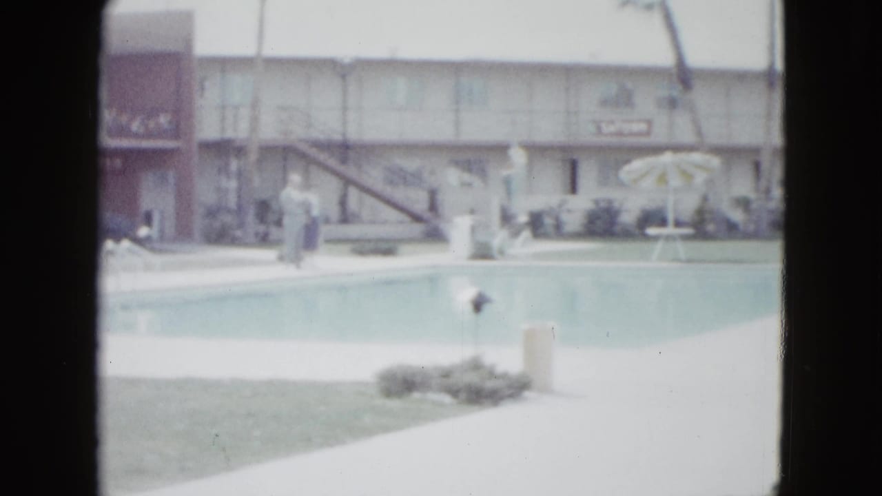 1958 Las Vegas home-movie frame showing a motel swimming pool, white deck, tan building, lawn, umbrella, and indistinct people.