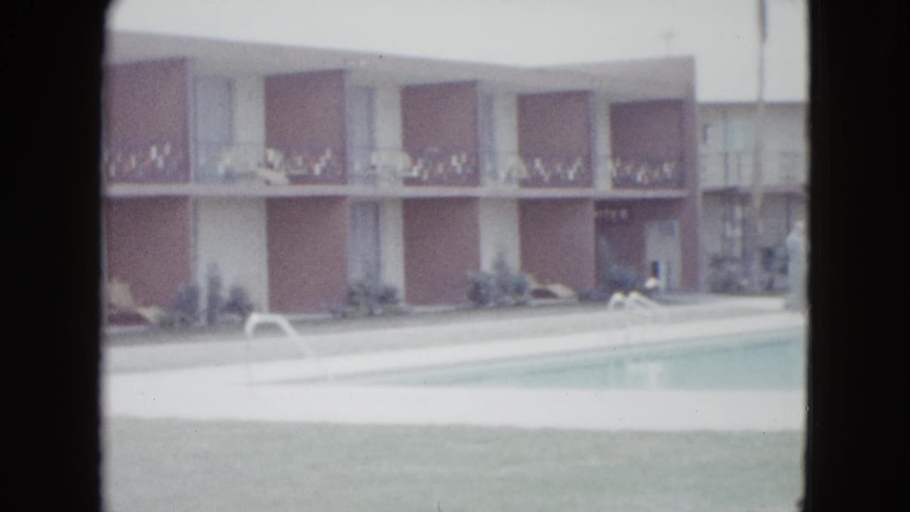 Soft 8mm frame of a 1958 Las Vegas motel pool with tan facade, balcony railings, shrubs, white deck, and lounge furniture.