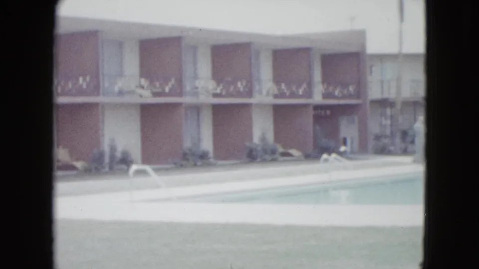 Soft 8mm frame of a 1958 Las Vegas motel pool with tan facade, balcony railings, shrubs, white deck, and lounge furniture.