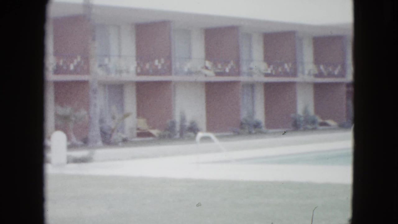 1958 Las Vegas motel courtyard view with pool water, white coping, lawn, shrubs, and a two-story building with repeated balcony bays.