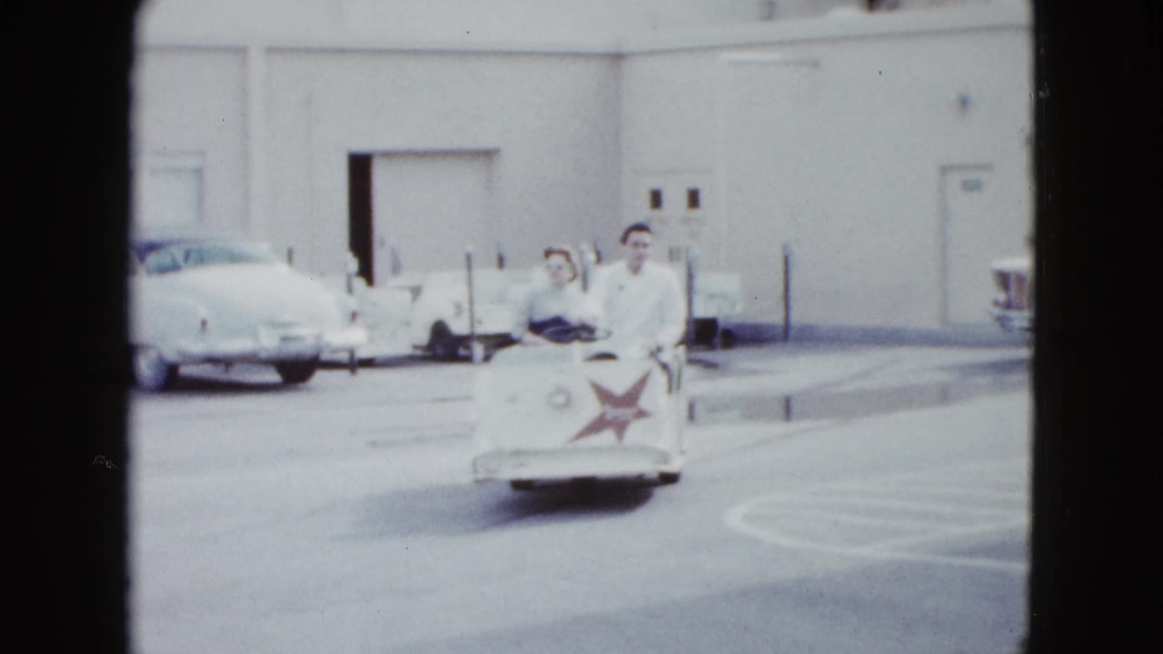 In Las Vegas, a couple rides a white hotel shuttle cart with a red star emblem past parked mid-century cars.