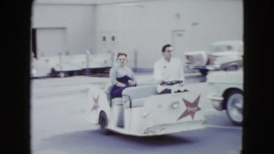 A woman passenger and male driver ride a white shuttle cart through a Las Vegas hotel parking area in 1958.