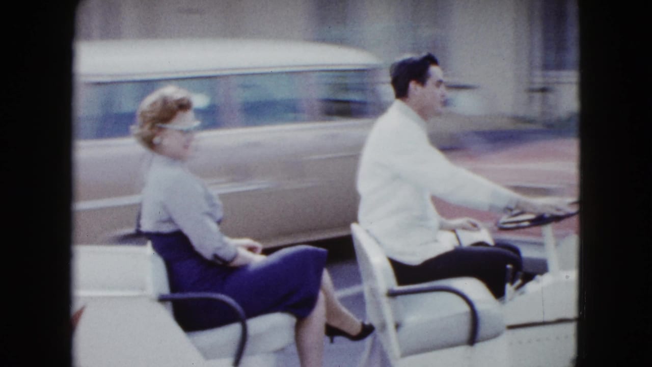 In 1958 Las Vegas footage, a couple rides a small white open car past motel buildings and parked period cars.