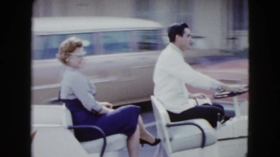 In 1958 Las Vegas footage, a couple rides a small white open car past motel buildings and parked period cars.