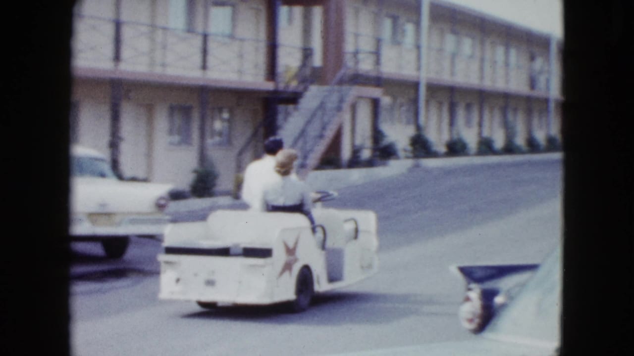 A rear view shows the couple riding away in the mini open car along a mid-century Las Vegas motel driveway.