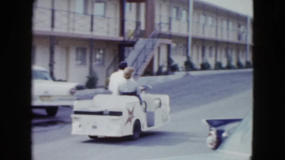 A rear view shows the couple riding away in the mini open car along a mid-century Las Vegas motel driveway.