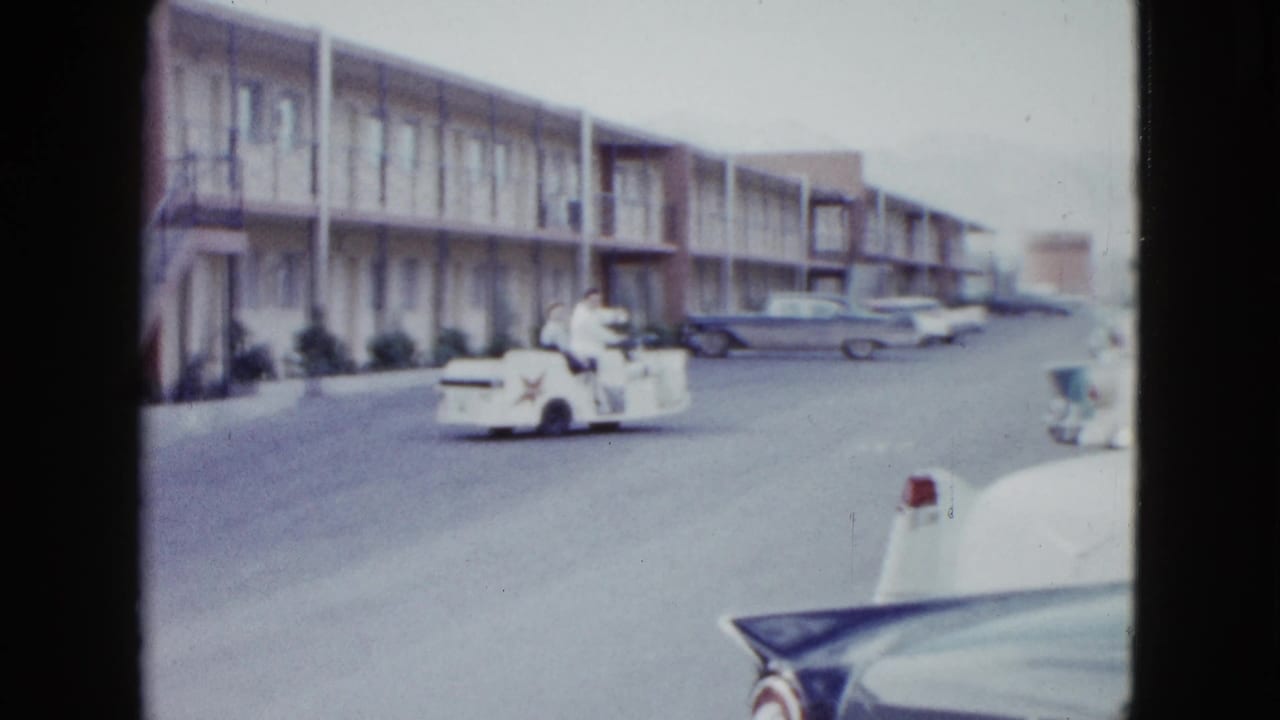 Cart with two people passes parked cars outside a long motel building.