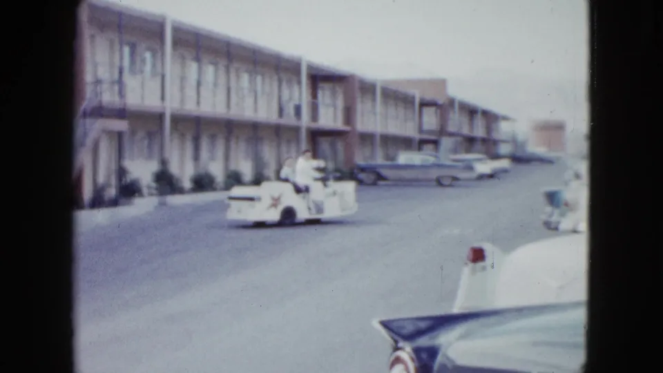 1958 Las Vegas frame with two people riding in a white cart along an exterior-corridor motel driveway.