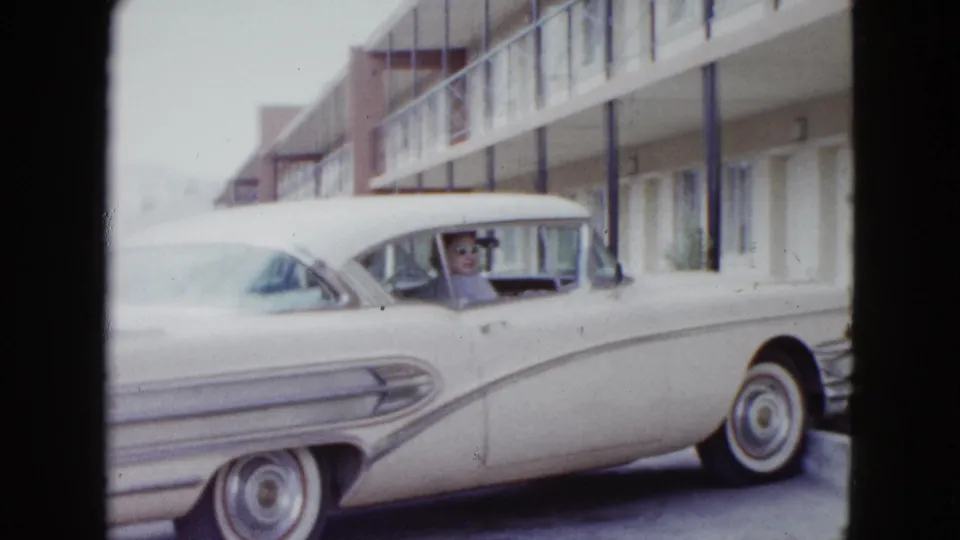 1958 Las Vegas home-movie frame showing a white classic sedan parked outside motel rooms, with a person visible through the window.