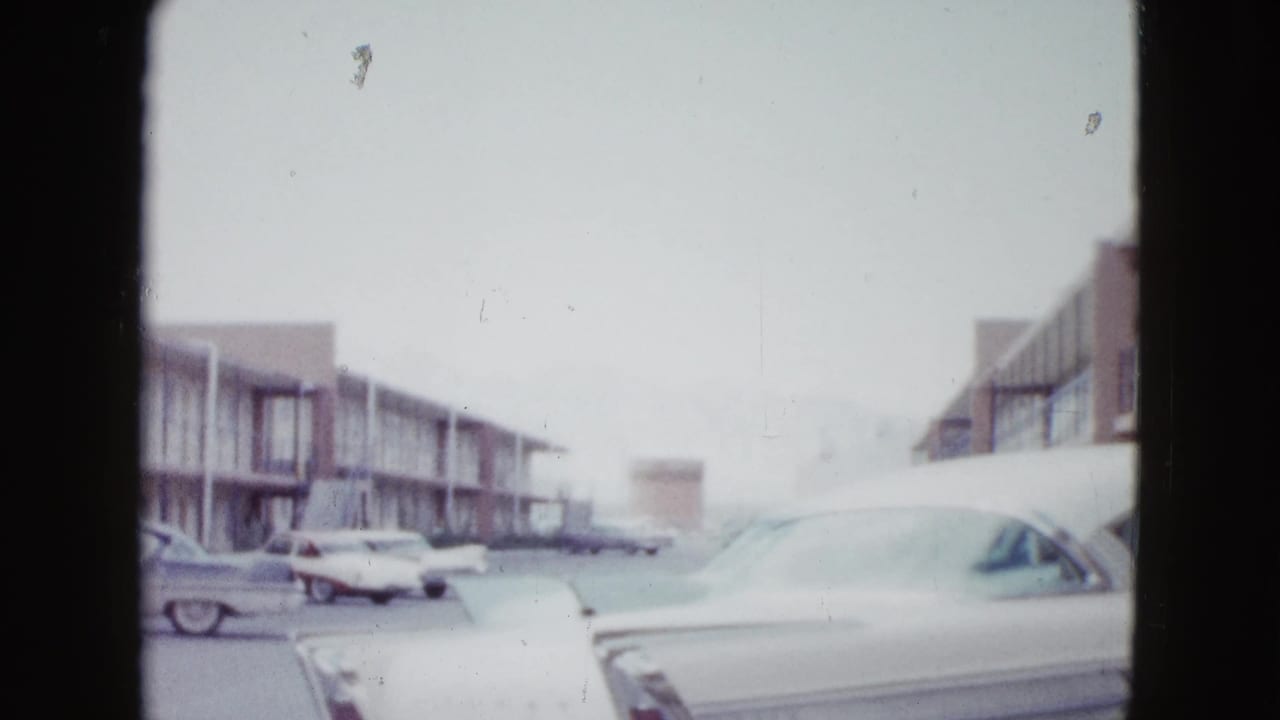 Motel parking lot with several vintage cars and a white sedan at right.