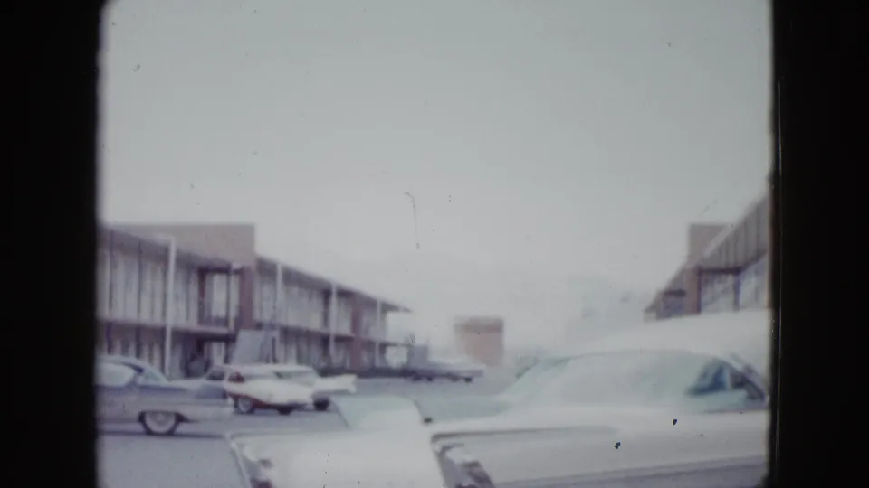 A soft 8mm frame shows parked mid-century cars beside a Las Vegas motel, with a large white car occupying the foreground.