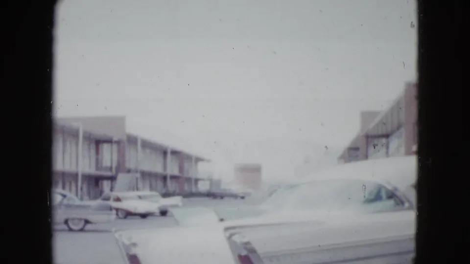 The closing frame holds on a white car beside a motel parking lot, with additional vintage cars and exterior balconies in the background.
