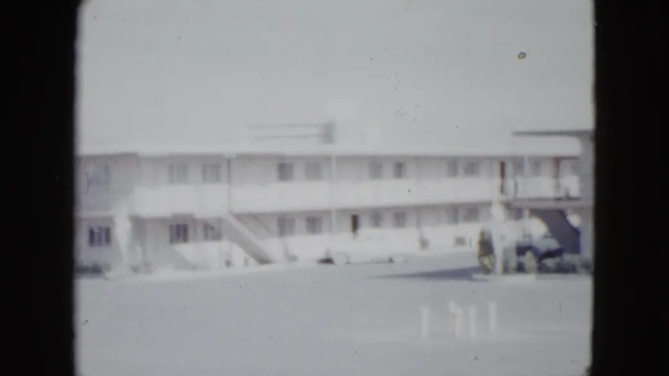 1958 Las Vegas motel exterior with repeating second-floor walkway, exterior stairs, paved lot, and a light-colored car near the right edge.