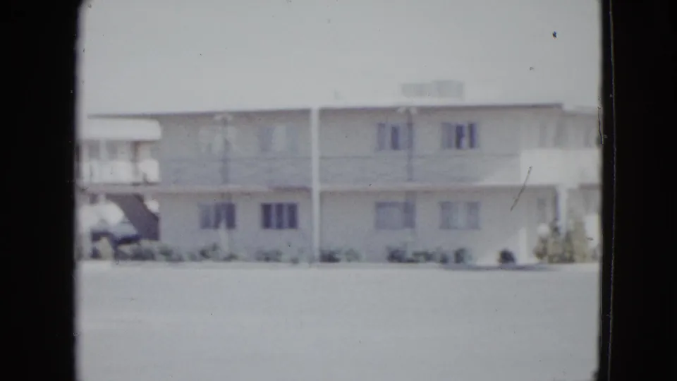 1958 Las Vegas home-movie frame of a motel side section with upper balcony, rectangular windows, low landscaping, and open paved foreground.