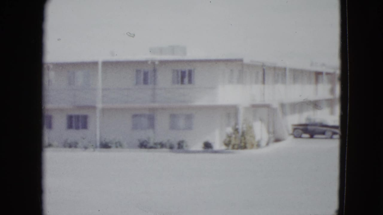 Sunlit two-story motel exterior with windows, exterior walkway, and a parked dark car.