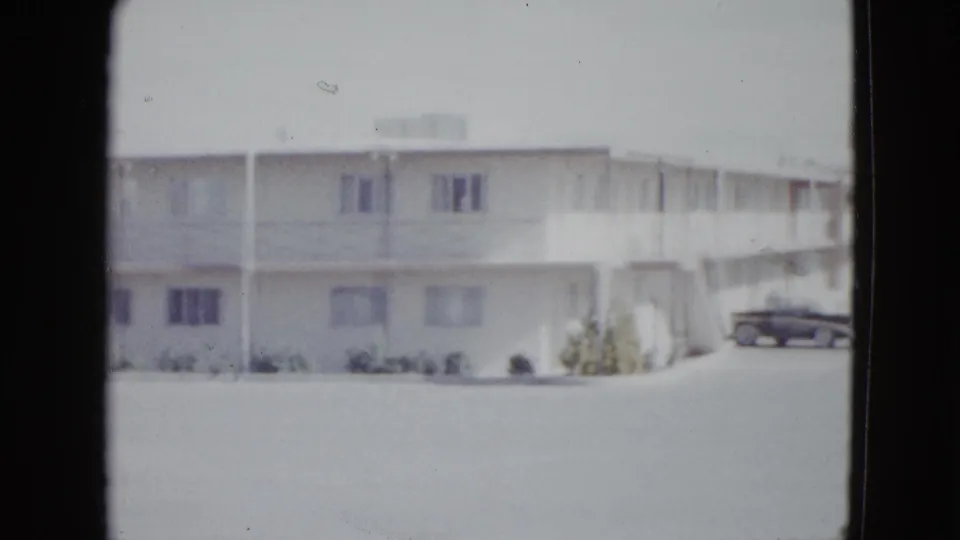 1958 Las Vegas home-movie frame showing a bright motel frontage with flat roof, windows, and a dark car parked near the building.
