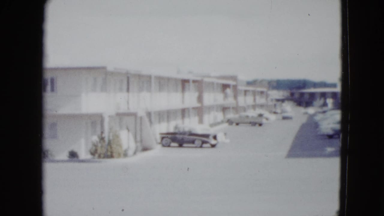 1958 Las Vegas frame with a two-story motel row, exterior corridors, parked late-1950s cars, and a bright open parking area.