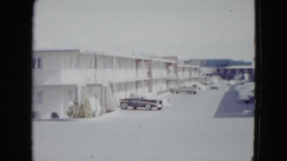1958 Las Vegas frame with a two-story motel row, exterior corridors, parked late-1950s cars, and a bright open parking area.