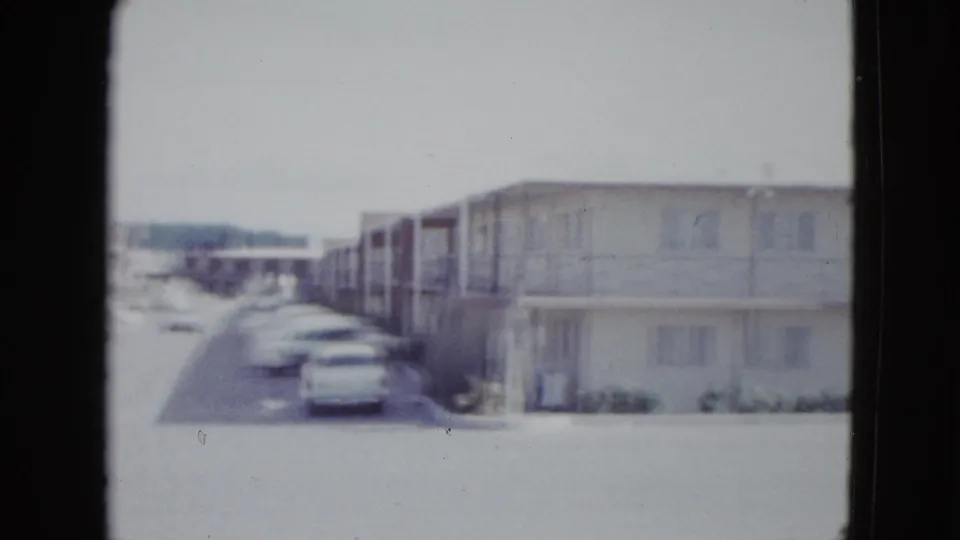 1958 Las Vegas frame showing the end of a two-story motel block, side parking, small shrubs, and cars under bright daylight.