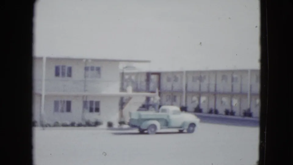 1958 Las Vegas home-movie frame with a light green pickup moving across a motel frontage with stairs and balcony railings.