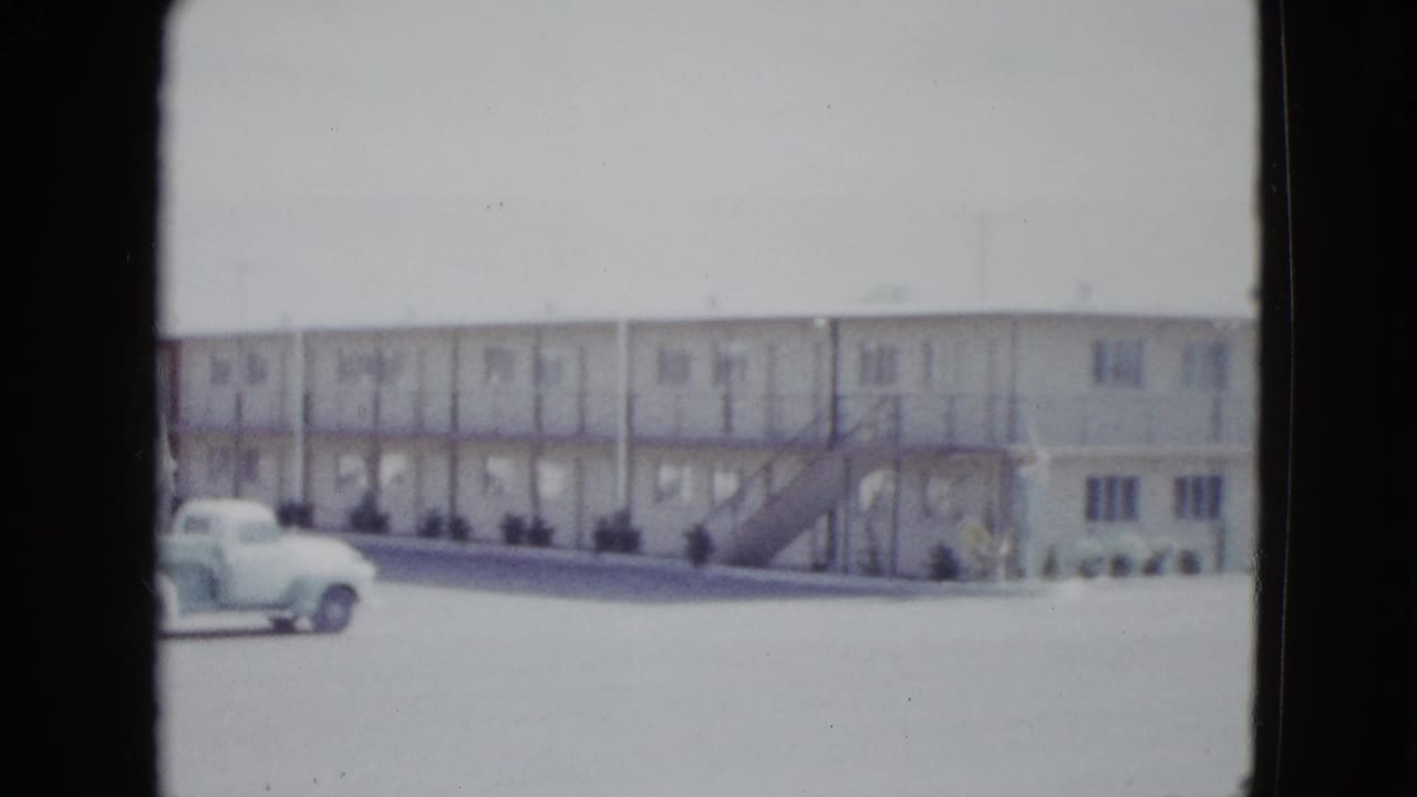 1958 Las Vegas home-movie frame showing a white two-story motel facade with exterior stairs and a light vintage pickup in front.