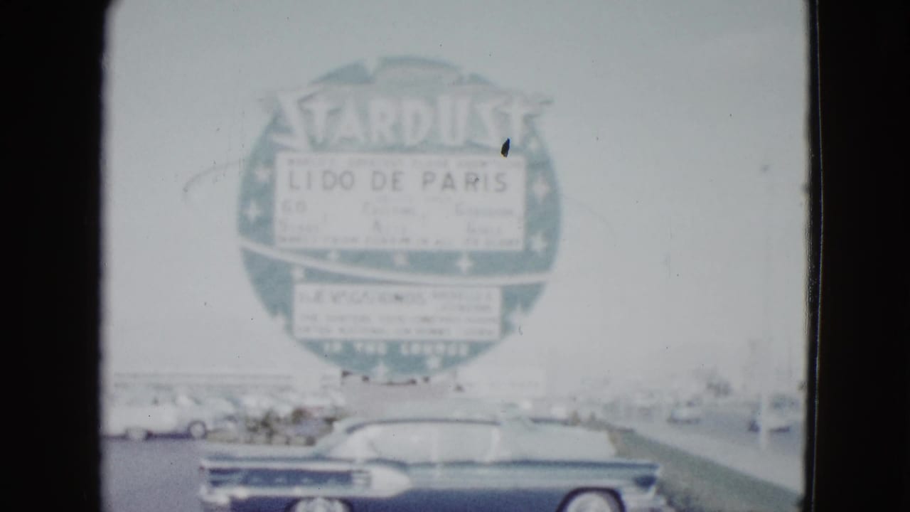Dark convertible below a large Stardust Lido de Paris sign on a bright street.