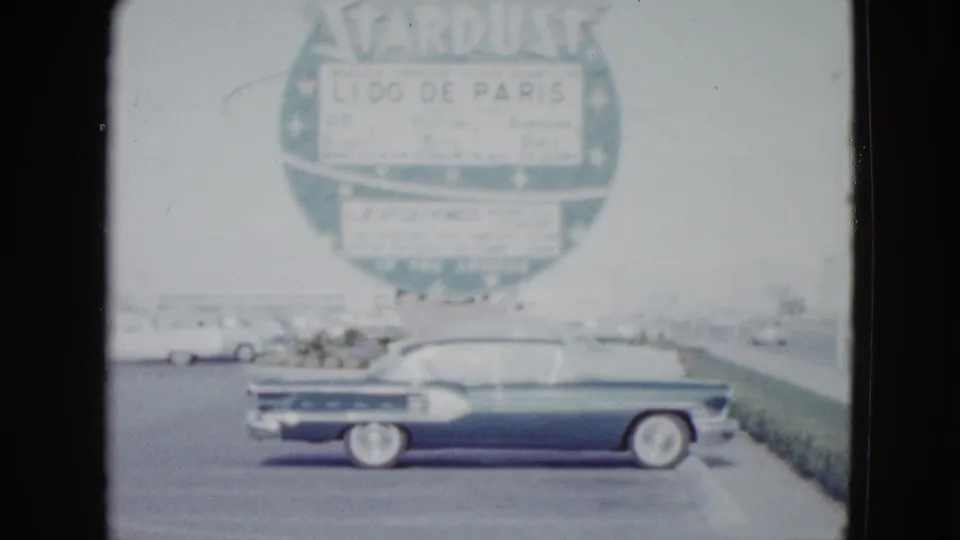 1958 Las Vegas view of a dark 1950s car near curb planting, with the Stardust Lido de Paris sign behind it.
