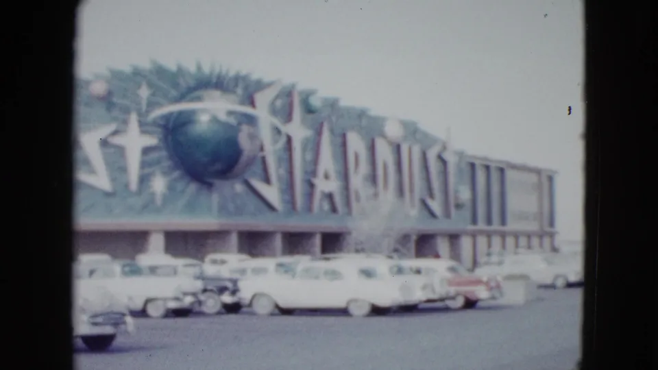 Closer handheld 8mm view of the Stardust Hotel sign and facade, with classic cars parked below in Las Vegas.