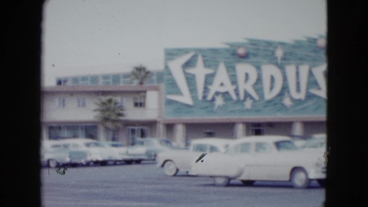 1958 Las Vegas frame from a left pan showing the Stardust sign edge, motel exterior, small palm, and parked classic cars.
