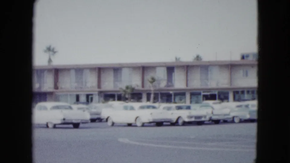 Wide Las Vegas parking-lot view showing mid-century cars, palm trees, and the hotel exterior near the end of the pan.