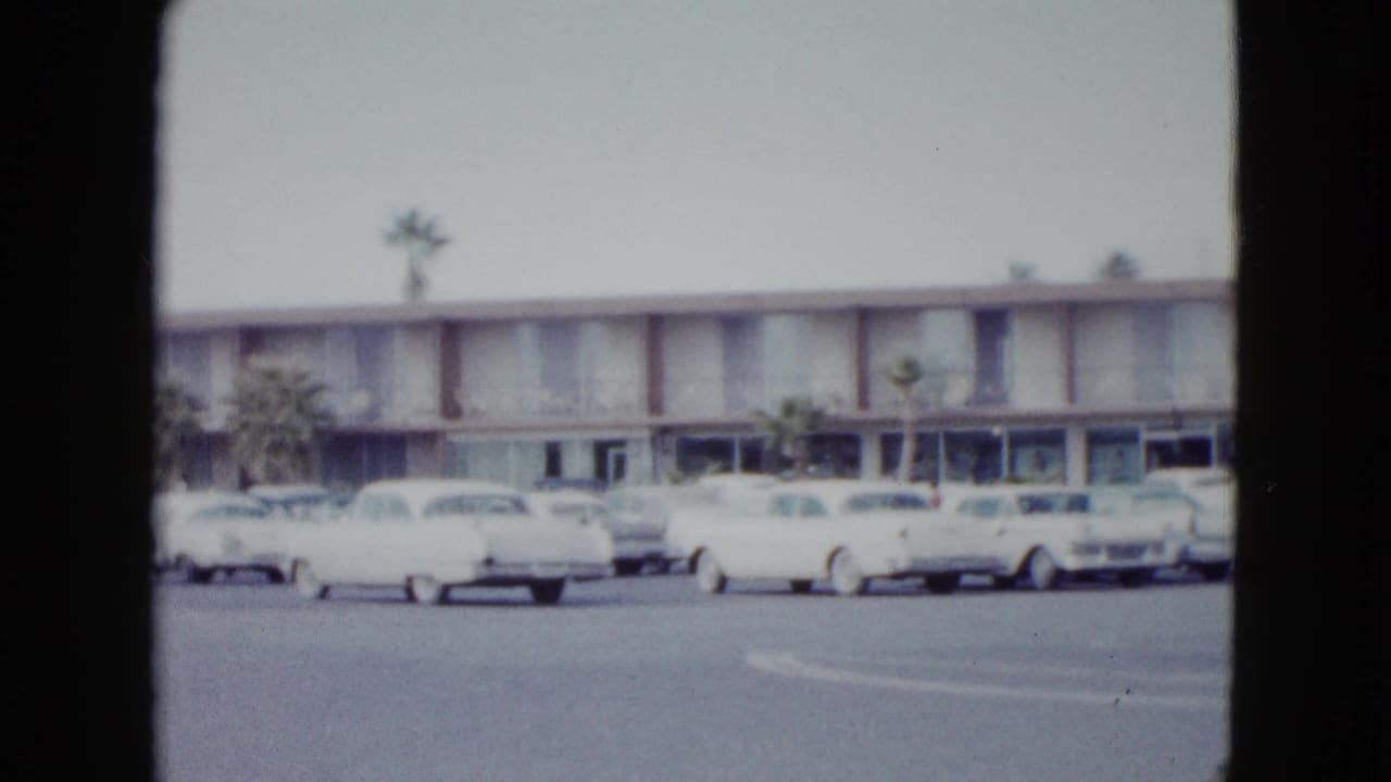 Two-story motel exterior with parked 1950s cars and palm trees across a lot.