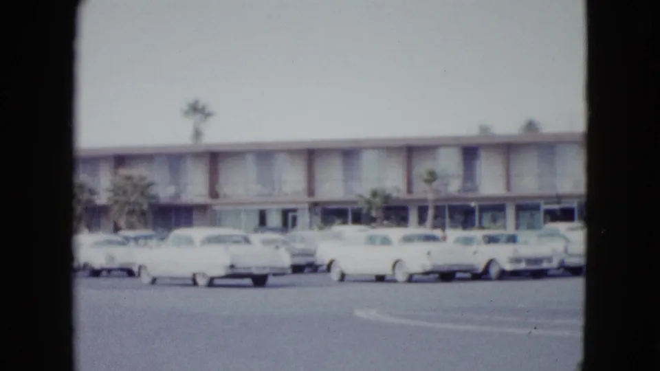 1958 Las Vegas 8mm frame showing a low motel exterior, palm trees, and several parked mid-century cars.